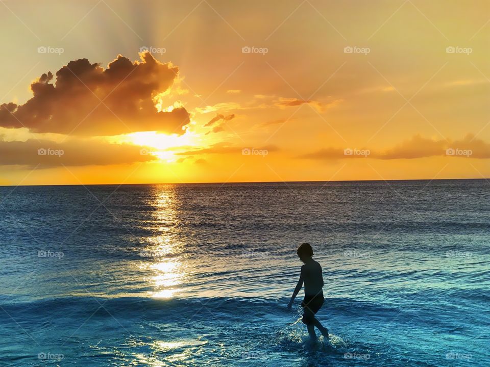 A young boy playing in the blue ocean waves during a glorious golden sunset.