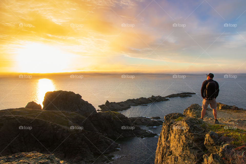 Young man, standing on an epic costal cliff watching a beautiful vibrant sunset. Newfoundland, Canada