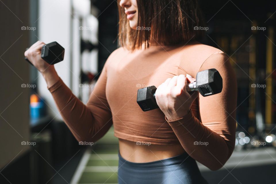 Young brunette woman training her muscles with dumbbells in the fitness club gym