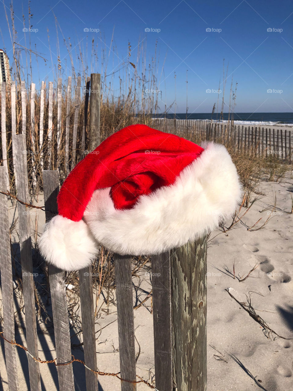 A Santa hat sits on a beach fence with the ocean in the background. 