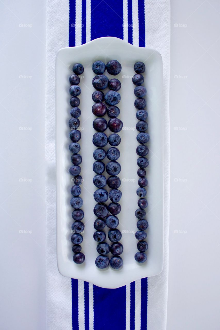 Vertical flat lay of blueberries on a rectangular white plate, arranged in lines to emphasize the blue stripes on the white dish towel underneath, all on a white background 