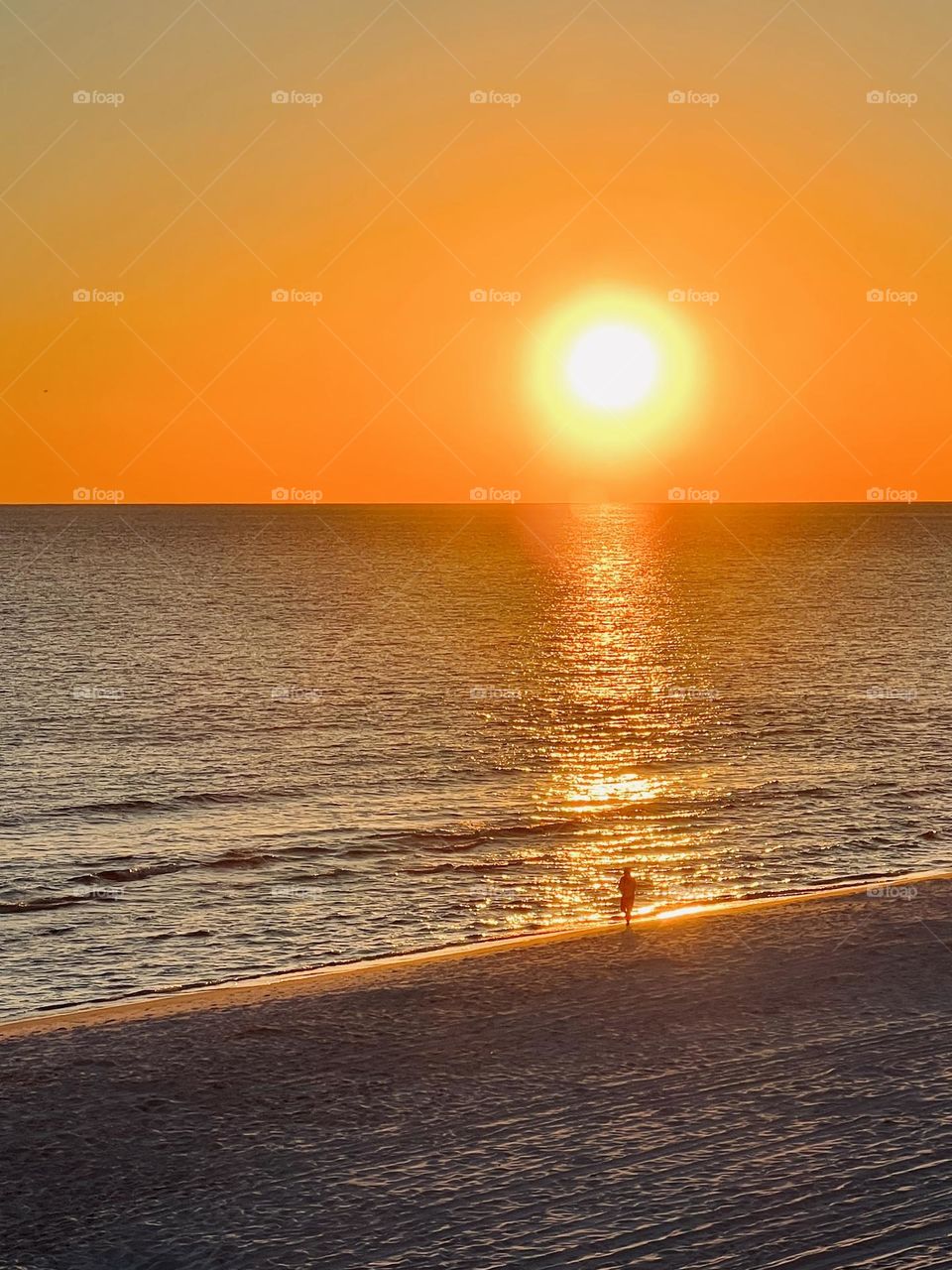 Running on the beach at sunset. A single person in silhouette running along the shoreline as the setting sun lends a orange hue to the water, sand and sky.