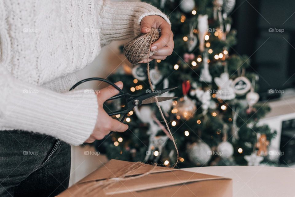 Midsection of woman wrapping christmas present in brown paper in front of christmas tree at home