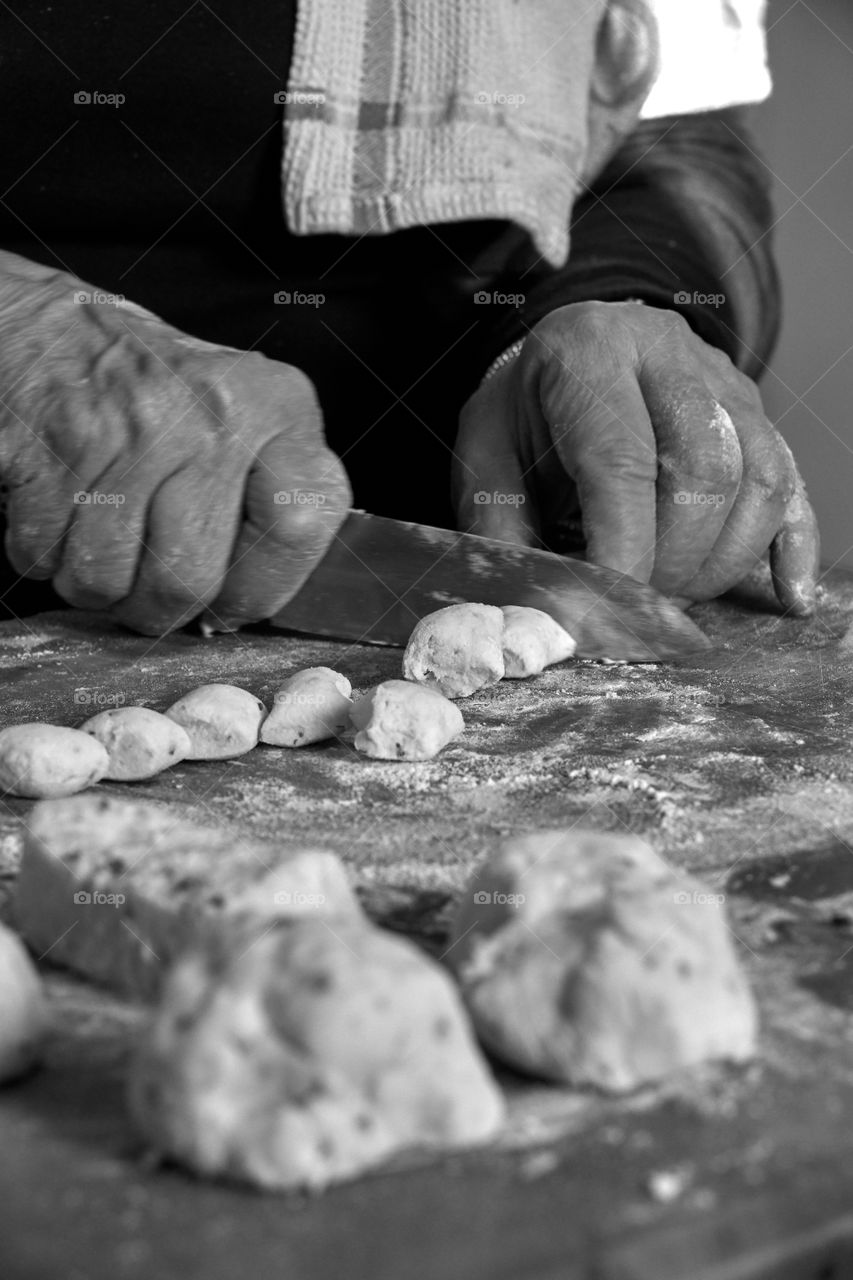 Monochromatic shot of old woman’s hands making and cutting fresh gnocchi in her kitchen on a floured butcher block. Shows skill, fresh ingredients, finely-honed skills, and cooking with love.