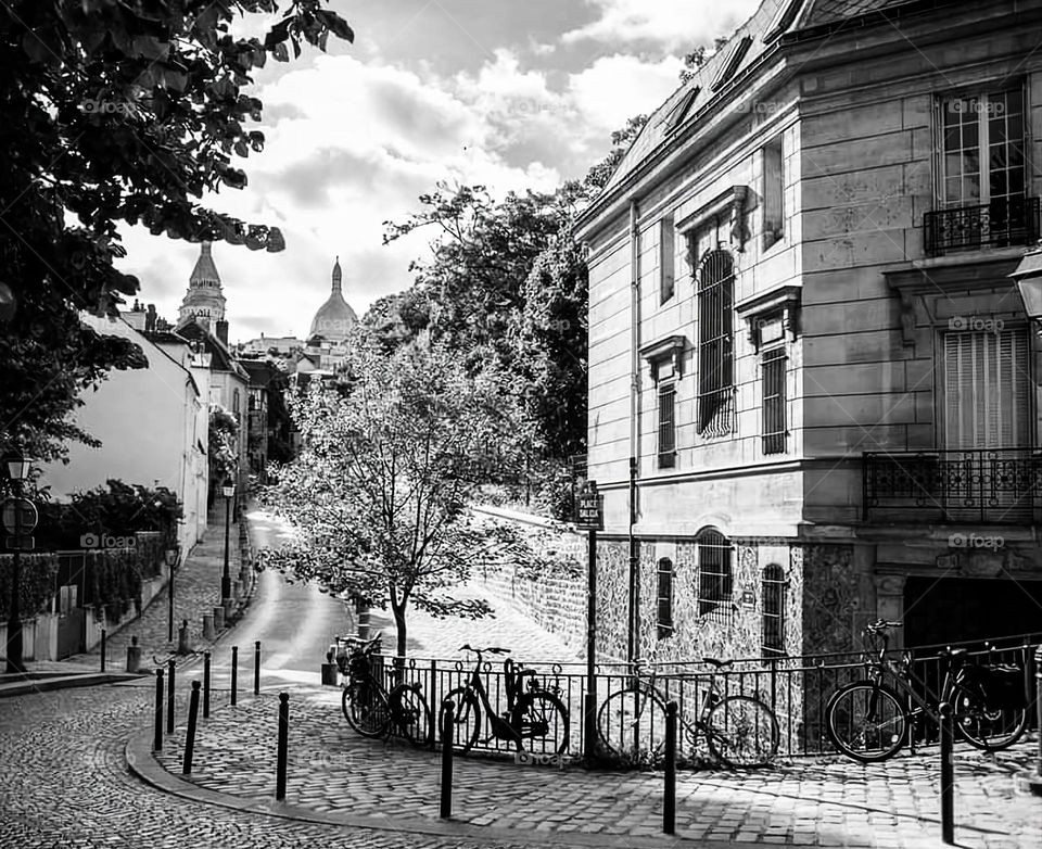 Vintage black and white shot of square Dalila in Montmartre with its trees, old stone building, paved street and bicycles along the wrought iron fence with a view of the Basilica of the Sacred Heart