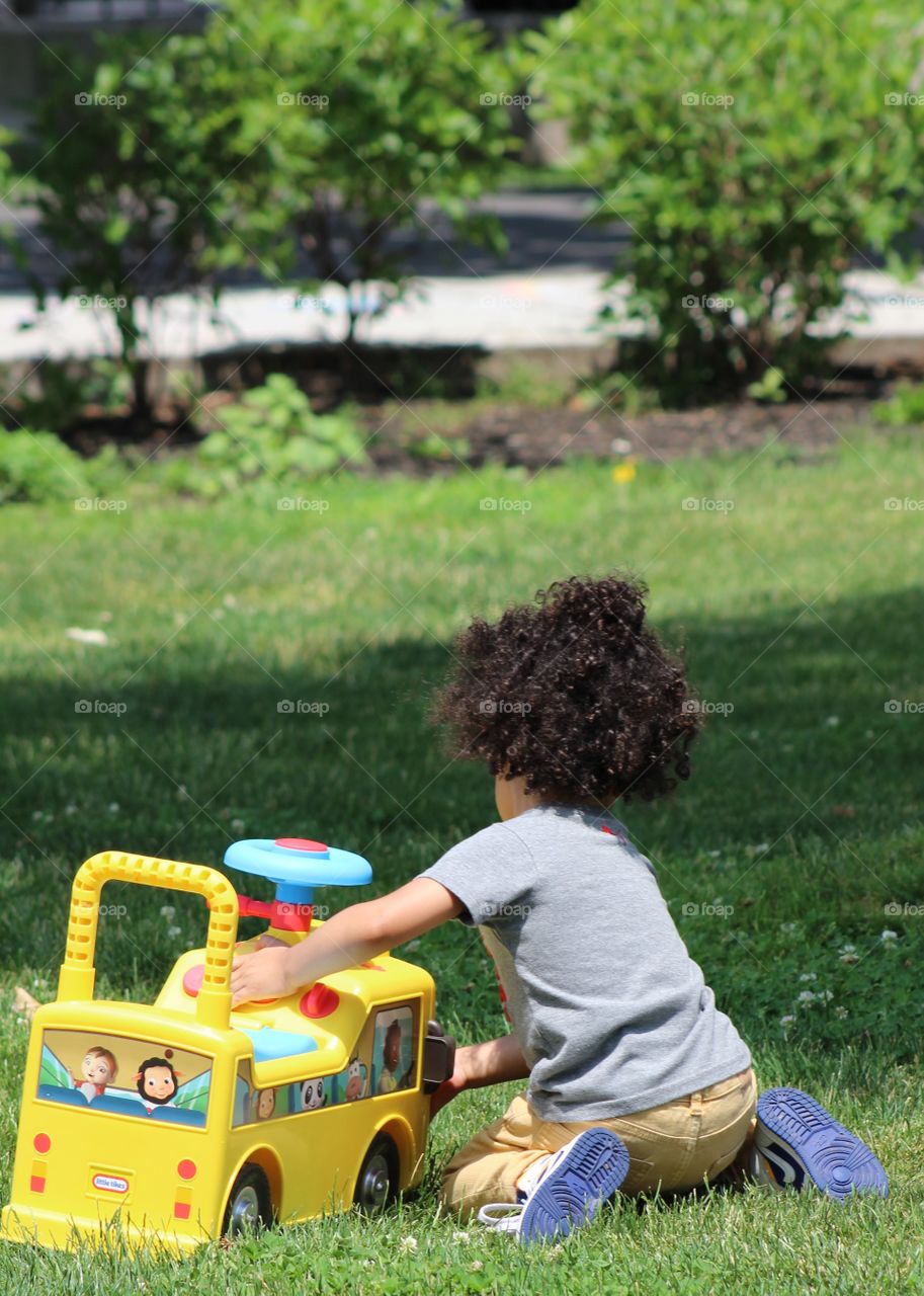 Toddler boy with Afro playing with yellow Little Tikes car in park in June 