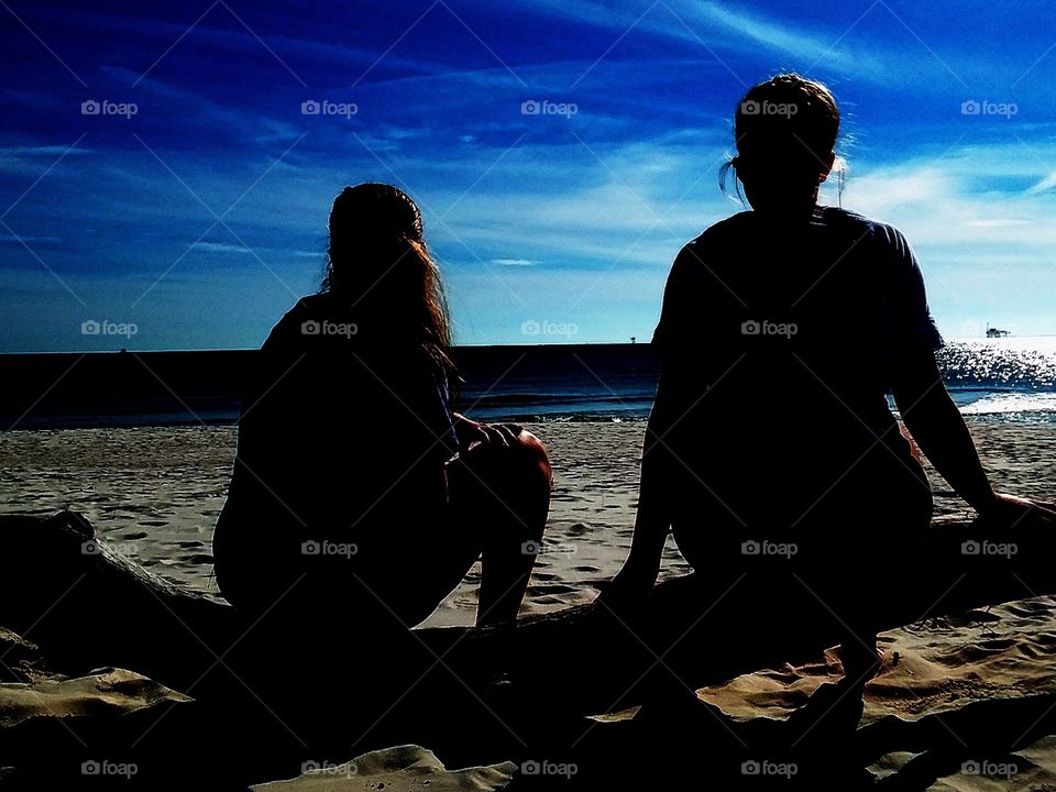 a pair of young girls sitting on a large piece of driftwood staring out into the ocean