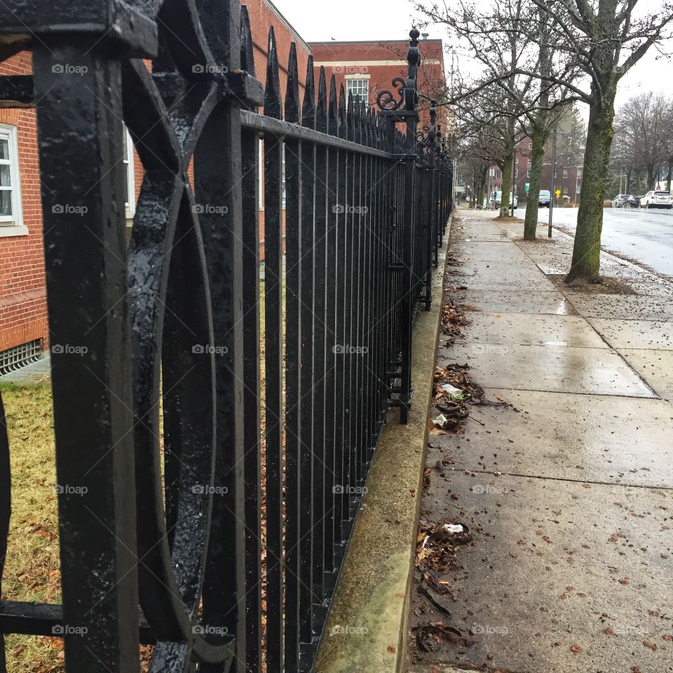 A wrought iron fence, painted black, as seen down it’s entire length. The top is made up of decorative points and there are curly decorations worked into each of the posts. The day is rainy.