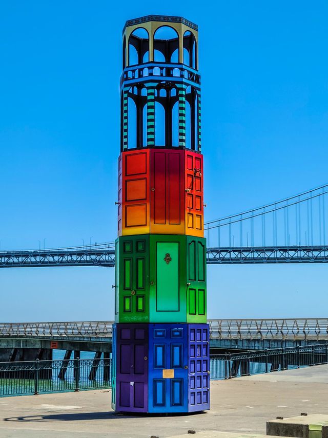 Art installation on the Embarcadero in San Francisco California in the shape of Coit Tower showing the doors of San Francisco in the rainbow colors of the Gay Pride Flag with tha Bay Bridge in the background 