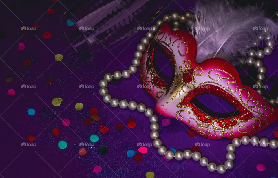 A beautiful pink masquerade mask with white feathers, pearl beads, colorful confetti and a champagne glass lie on a lilac background, close-up side view. Masquerade accessories concept.