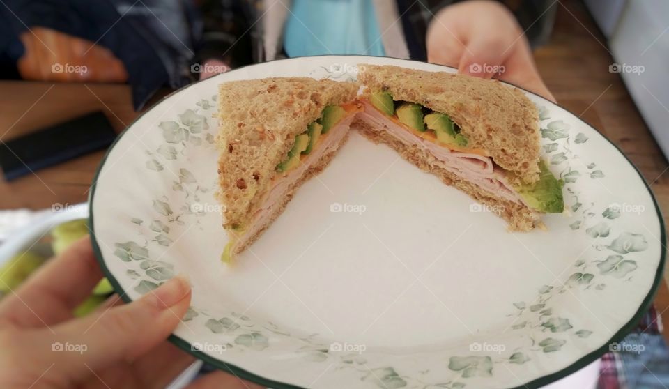 A woman's hand handing a man's hand a plate with his favorite sandwich a crust less turkey cheese avocado dijon mustard on whole grain wheat bread
