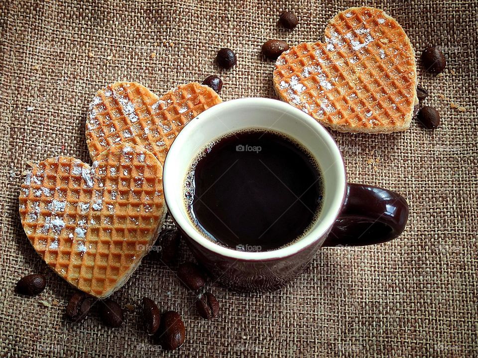 On the canvas is a brown mug with black coffee.  Around the cup are waffles in the shape of a heart and grains of black coffee