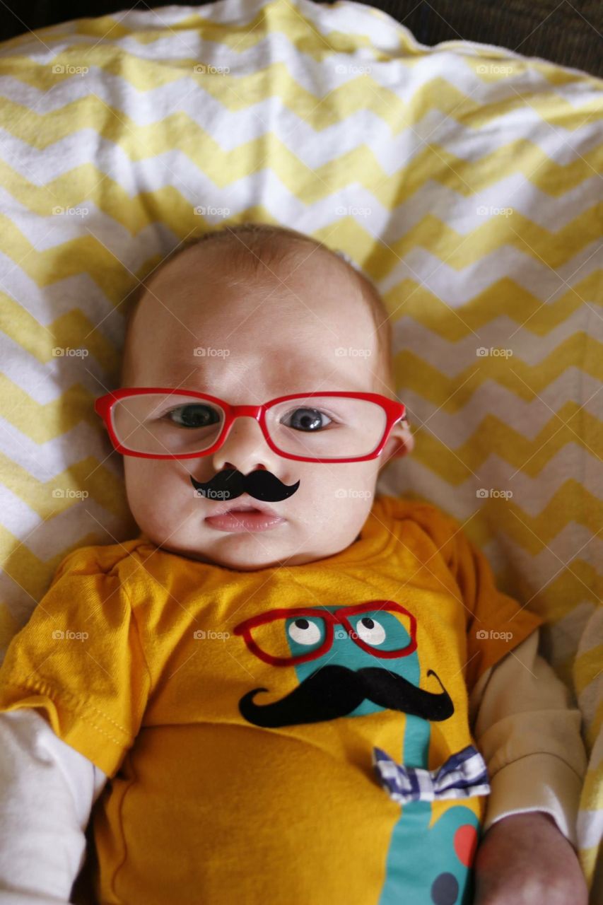 Red glasses. A baby wears a shirt with a dinosaur wearing red glasses while wearing red glasses