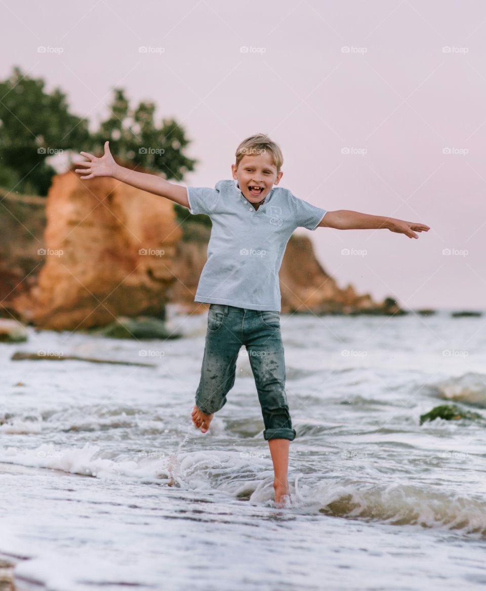 Joyful boy runs along the seashore spreading his arms like wings