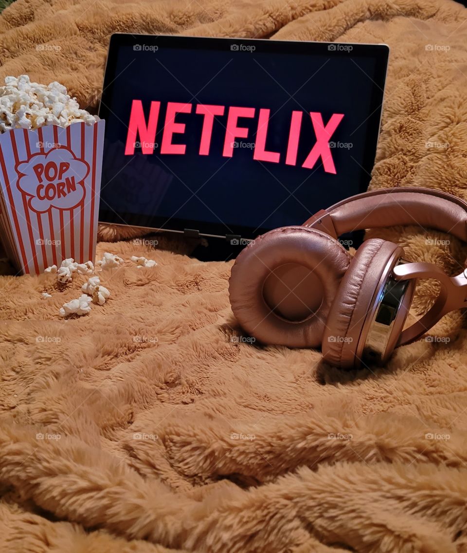 Laptop computer screen ready to watch Netflix for a fun, cozy, and relaxing night with popcorn as a snack, on soft bed blanket in dark room, with pink headphones