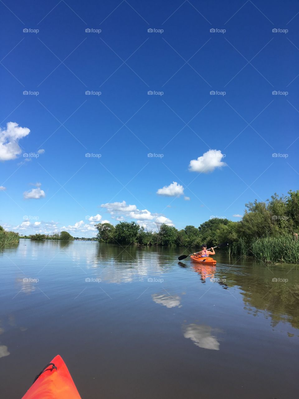 Woman kayaking on a smooth channel with green vegetation, blue skies & puffy white clouds.