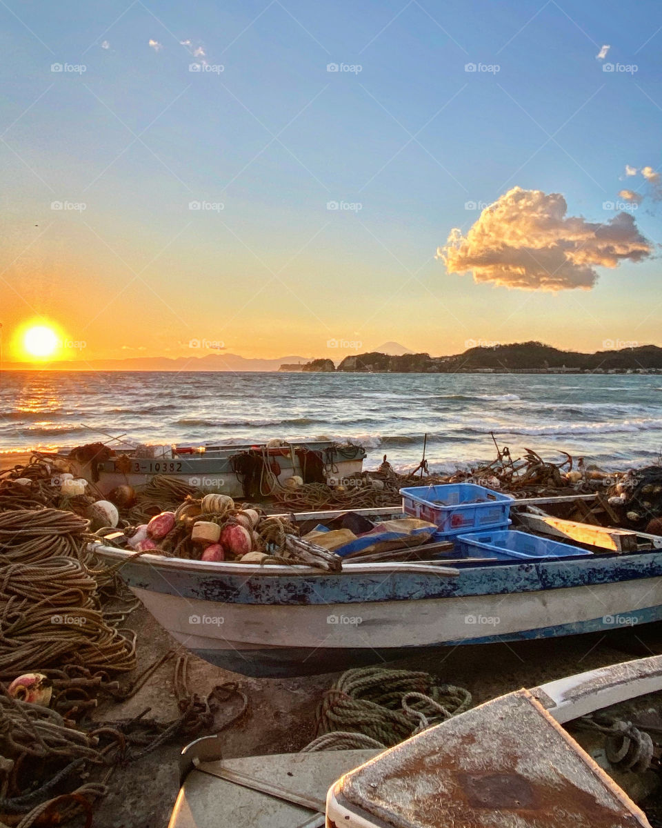 Golden hour sun setting into a sea with choppy waves, airplane shaped cloud floating in an otherwise clear sky, fishing boats, with ropes, crates and buoys in the foreground.