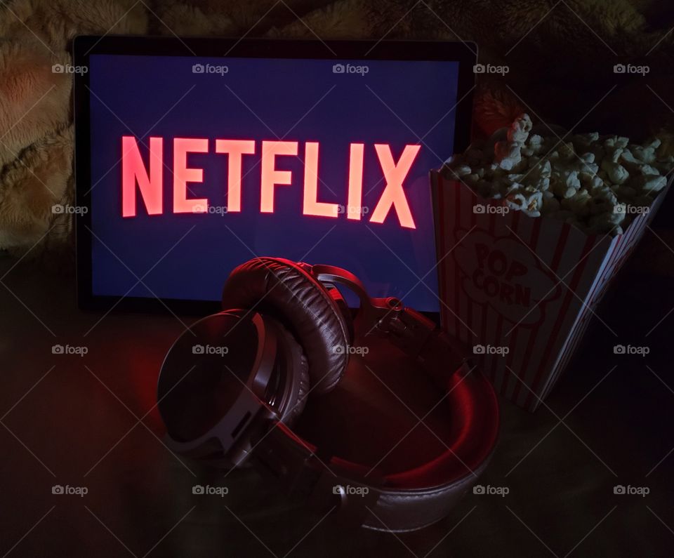 Neon letters display on laptop computer screen ready to watch Netflix for a fun, cozy, and relaxing night with popcorn as a snack, on soft bed blanket in room, with pink headphones