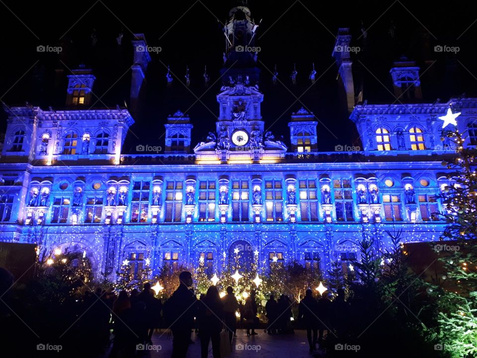 Vast classic building with Christmas lights and crowd gathering by night in front of Christmas trees