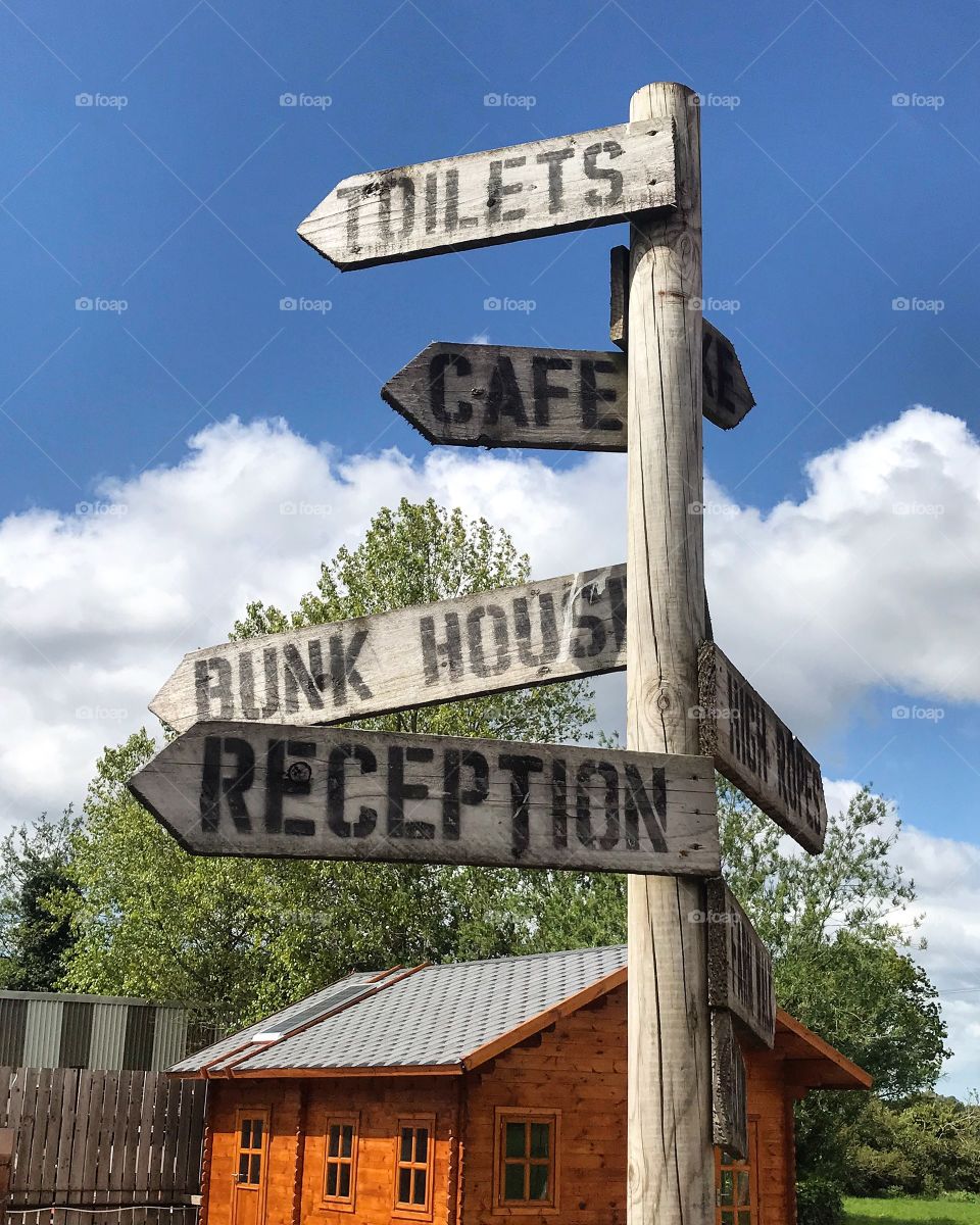 Taken at a child’s camp, the image is of a directional sign made of wood, against a clear and beautiful blue sky 