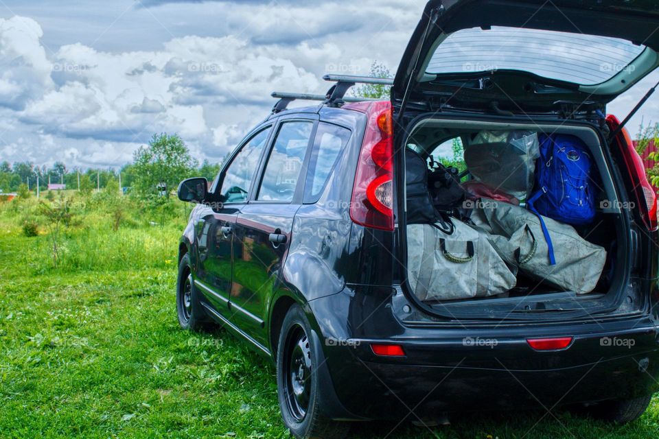 the black nissan note is stuffed full of bags with motors, boats, fishing rods and other things for river rafting. open trunk Green grass and clouds in the sky