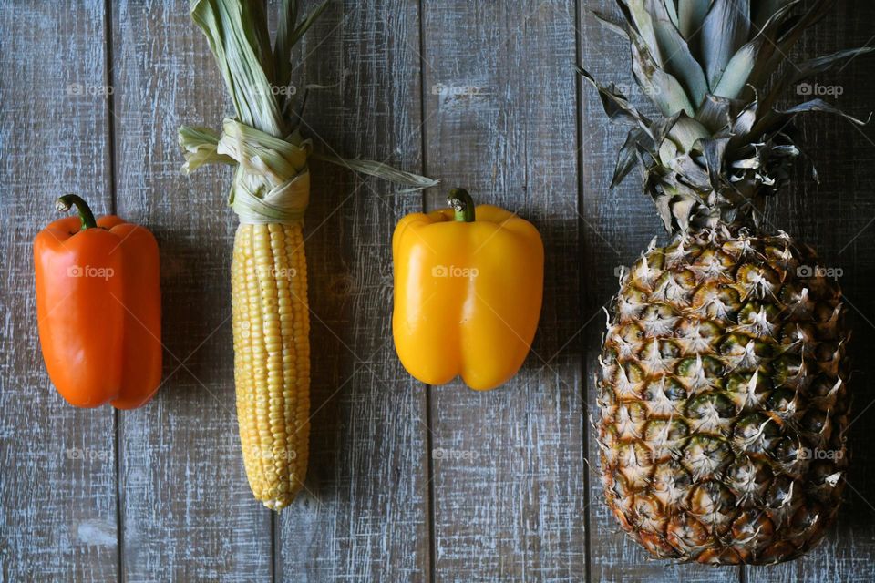 A pineapple, 2 bell peppers, and a corn on the cob lay out on a wooden cutting board.