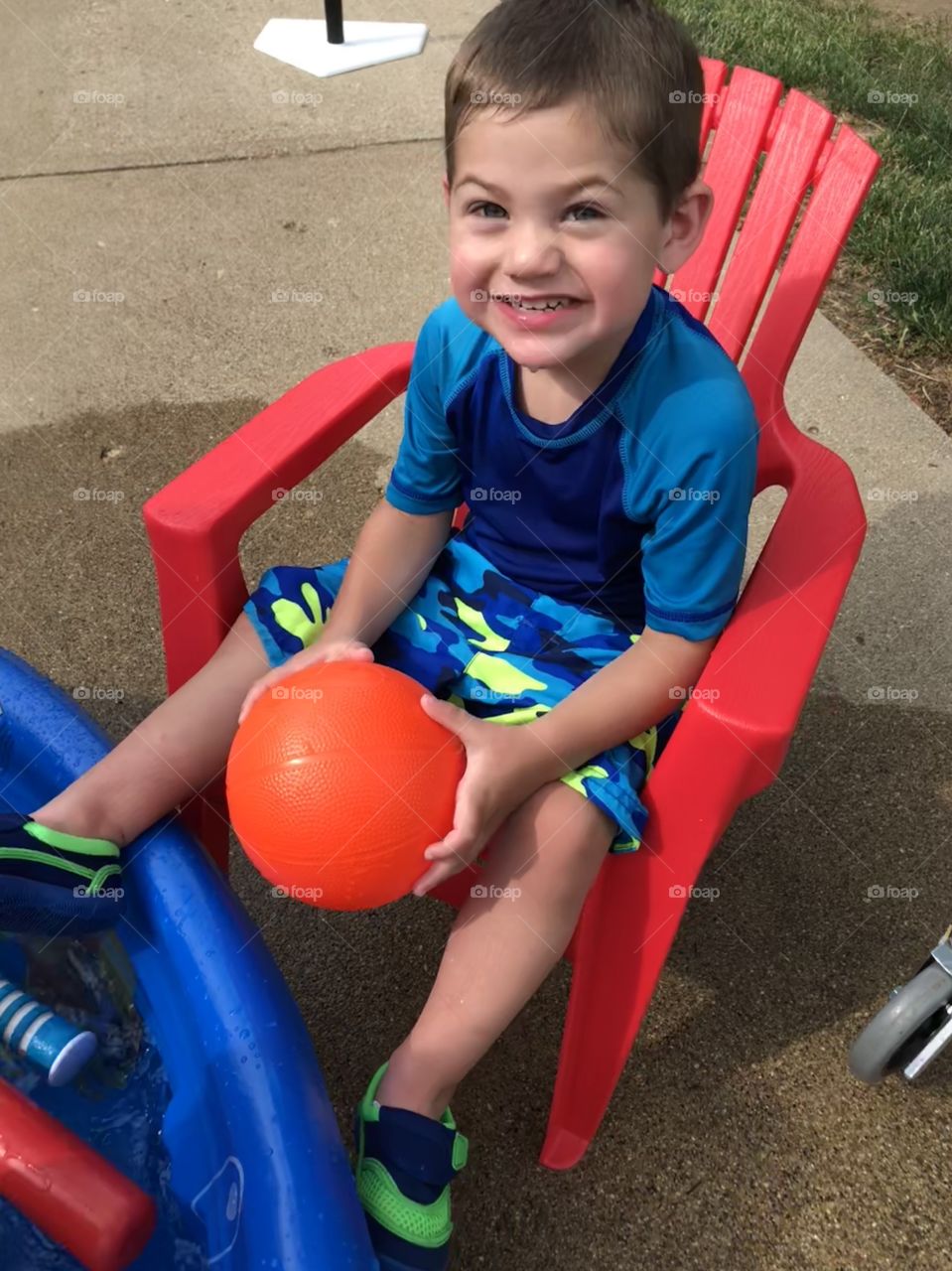 Toddler boy in swim trunks sitting by small pool holding a ball