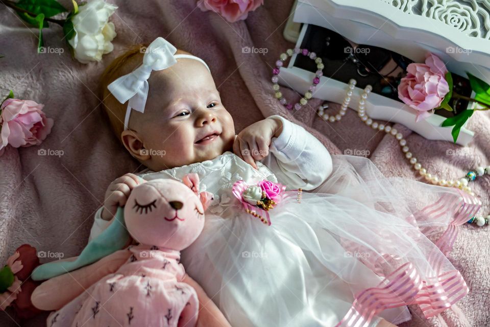 Little cute girl in a doll dress lies on a pink bed with beads and a plush hare and smiles