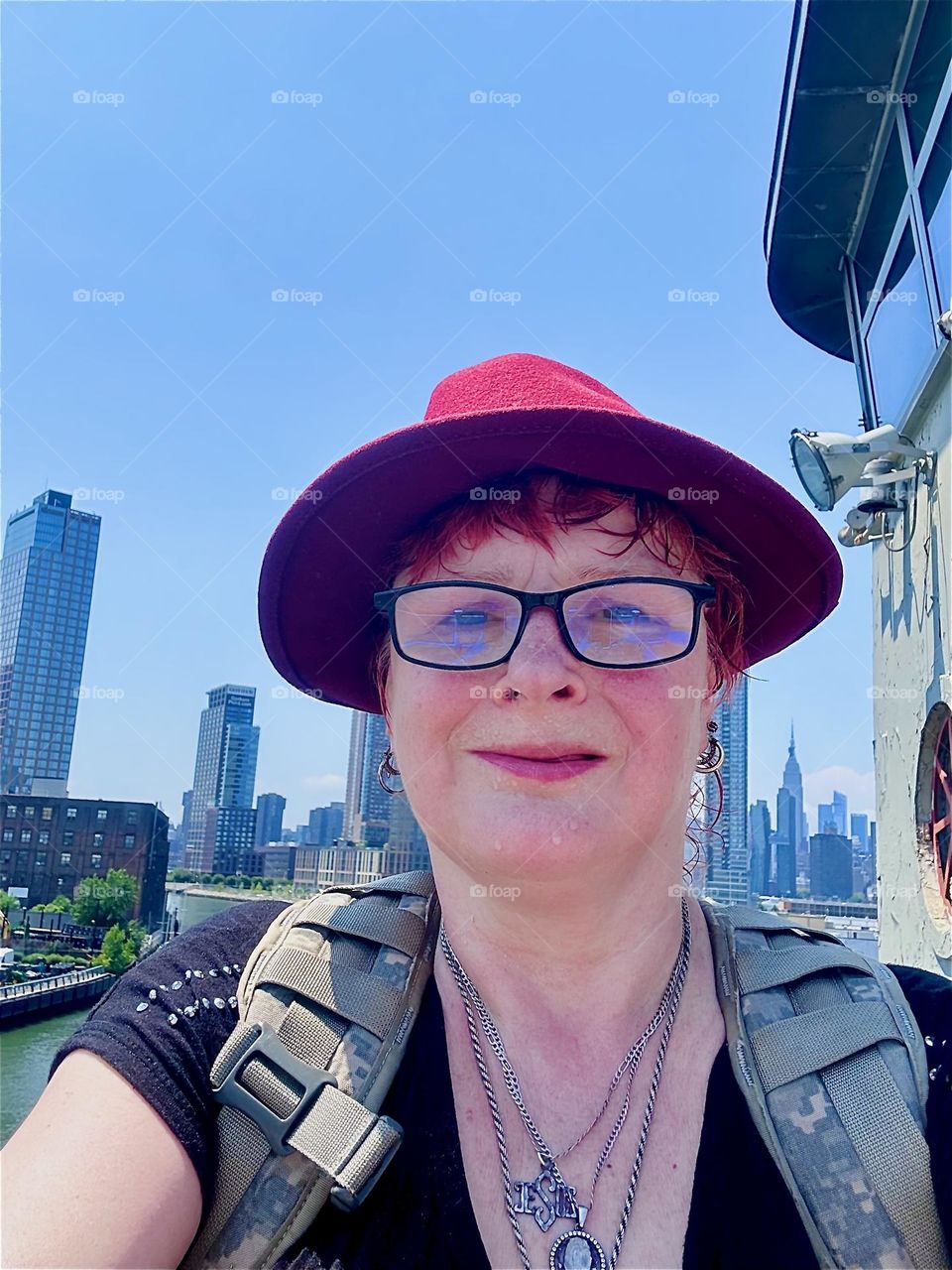 I, “Petra Schiller” a.k.a. “Maxine Firefox” am standing by the outpost of the “Pulaski Bridge” at “Newtown Creek” that connects LIC to “Greenpoint”, Bklyn on a midsummer day with “Manhattan”, LIC and “Greenpoint” behind me. 2024. Hypnotic Productions