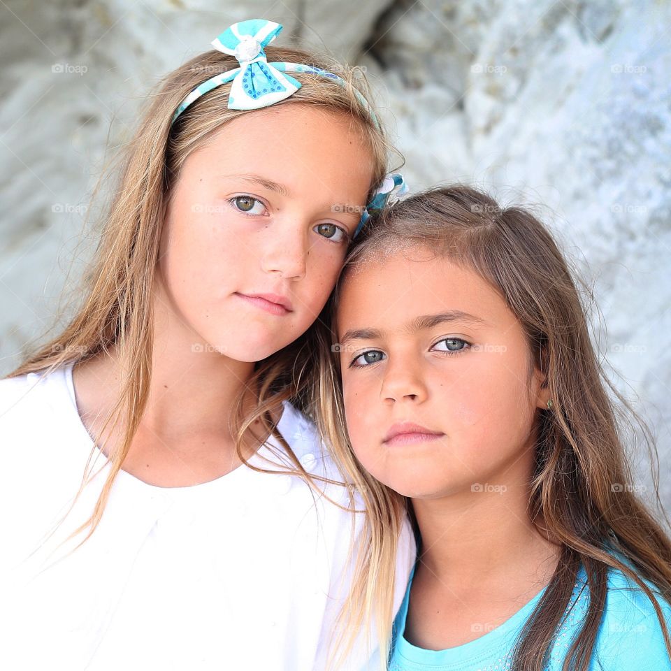 Outdoor beach portrait of two young sisters girls with serious pleasant expressions long hair headband bow with white and teal outfits
