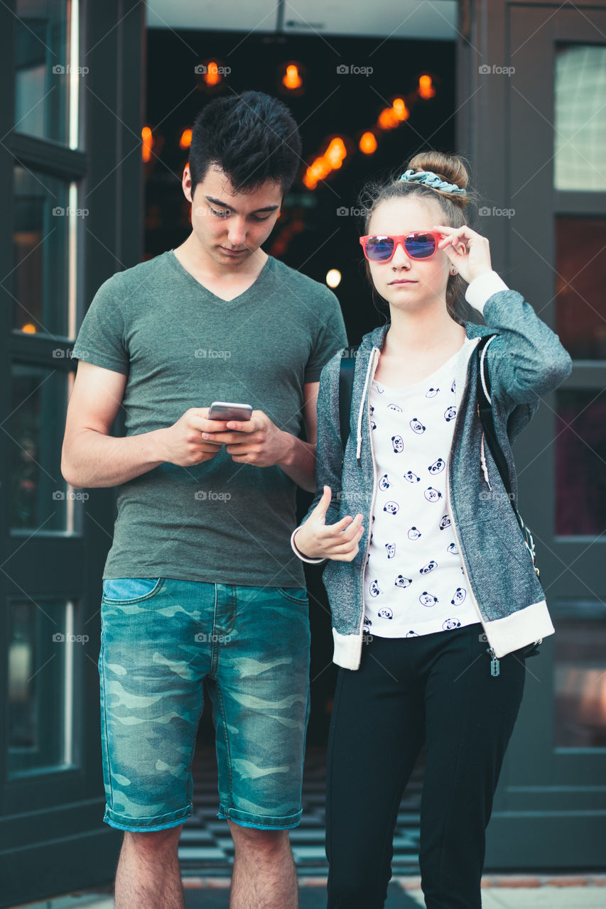 Couple of friends, teenage girl and boy, using smartphones, talking together, standing on street in center of town, spending time together