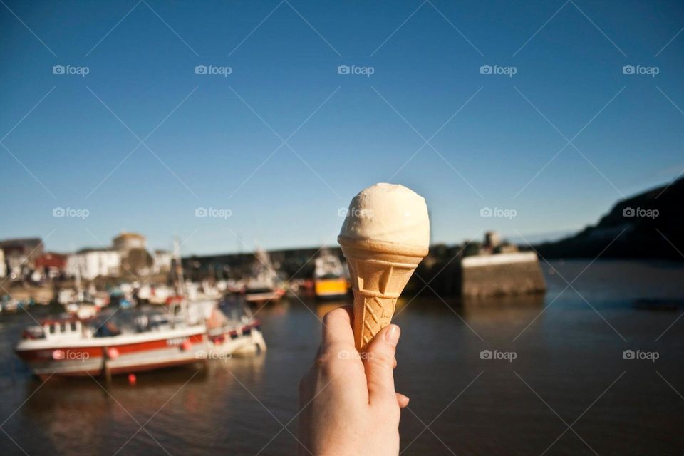 An outstretched hand holds an ice cream cone in front of blue sea & sky