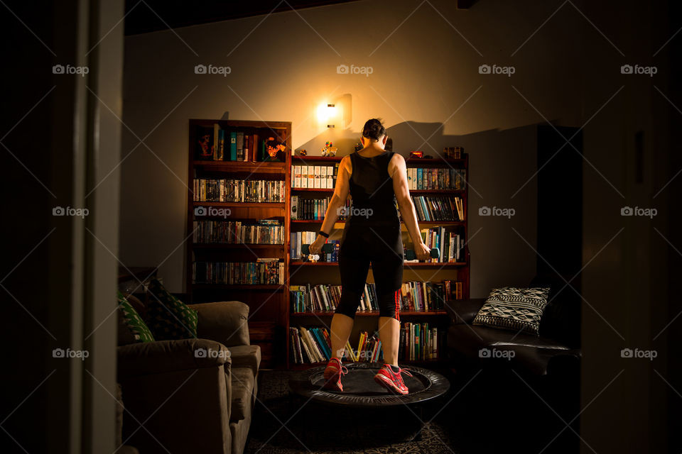 Staying fit and in shape at home during Covid lockdown. Image of woman in lounge with bookshelf training on small trampoline with dumbells at night.