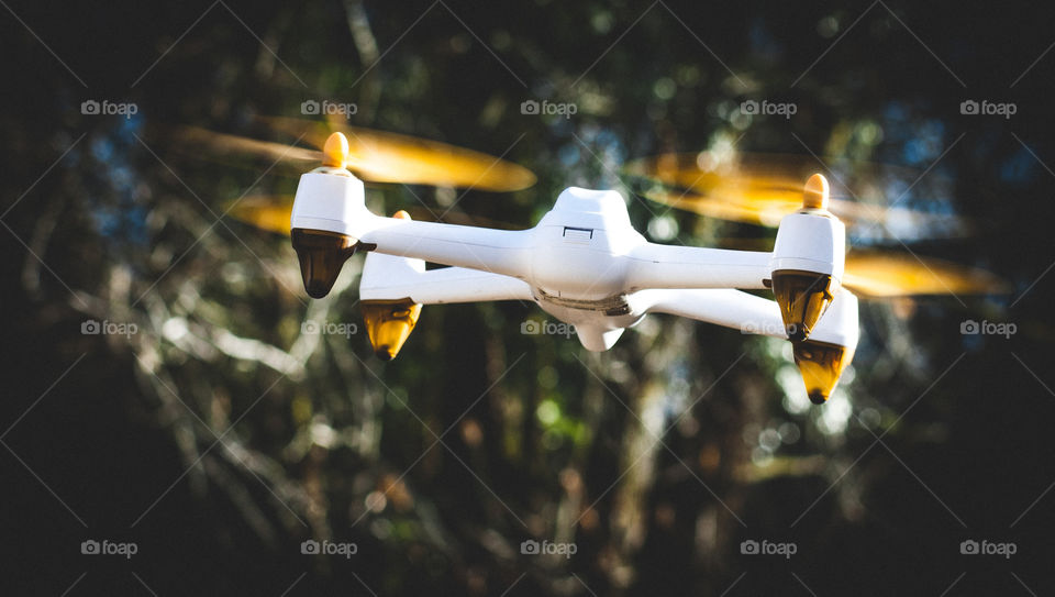 A white drone, hoovers with it propellers spinning, against a dark background of foliage
