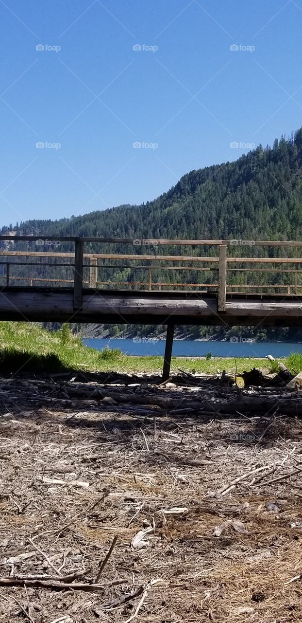 a wooden foot bridge, green grass with view of lake and mountain ridge under a sunny blue sky
