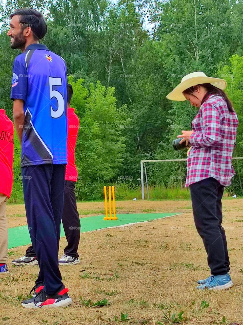 Summer day. A field surrounded by green trees. A girl in a straw hat is standing on the field and looking into the photo camera at the footage. Nearby are stand men who play cricket