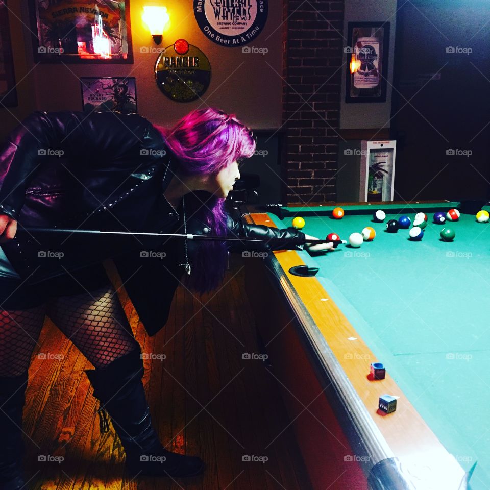 A woman with purple hair, fish net tights and a leather jackets shooting a game of pool.