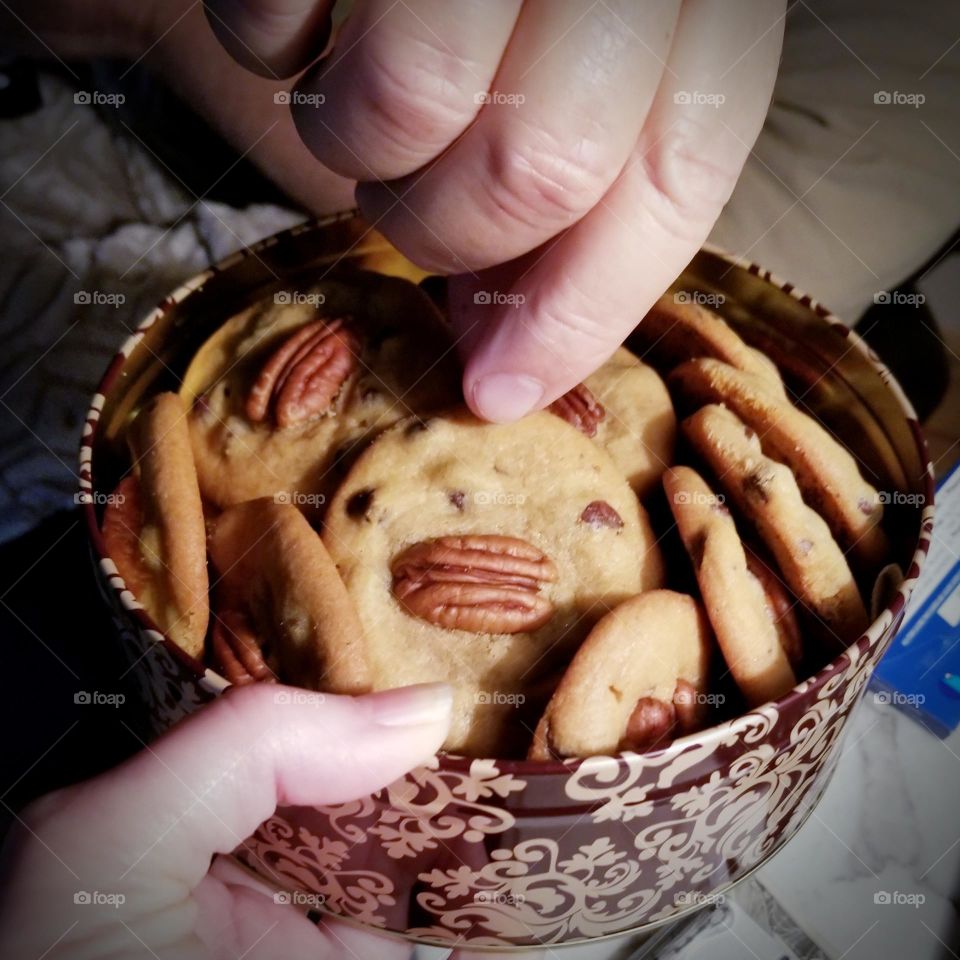 A woman holding a tin of Chocolate Chip Pecan Cookies while a man reaches for one