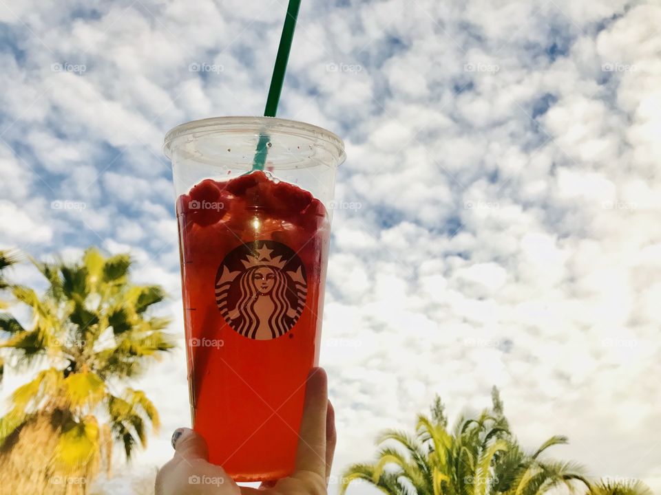 A woman holding a Starbucks pink strawberry Teavana drink high up against the cloudy blue sky and green tropical palm trees. USA, America 