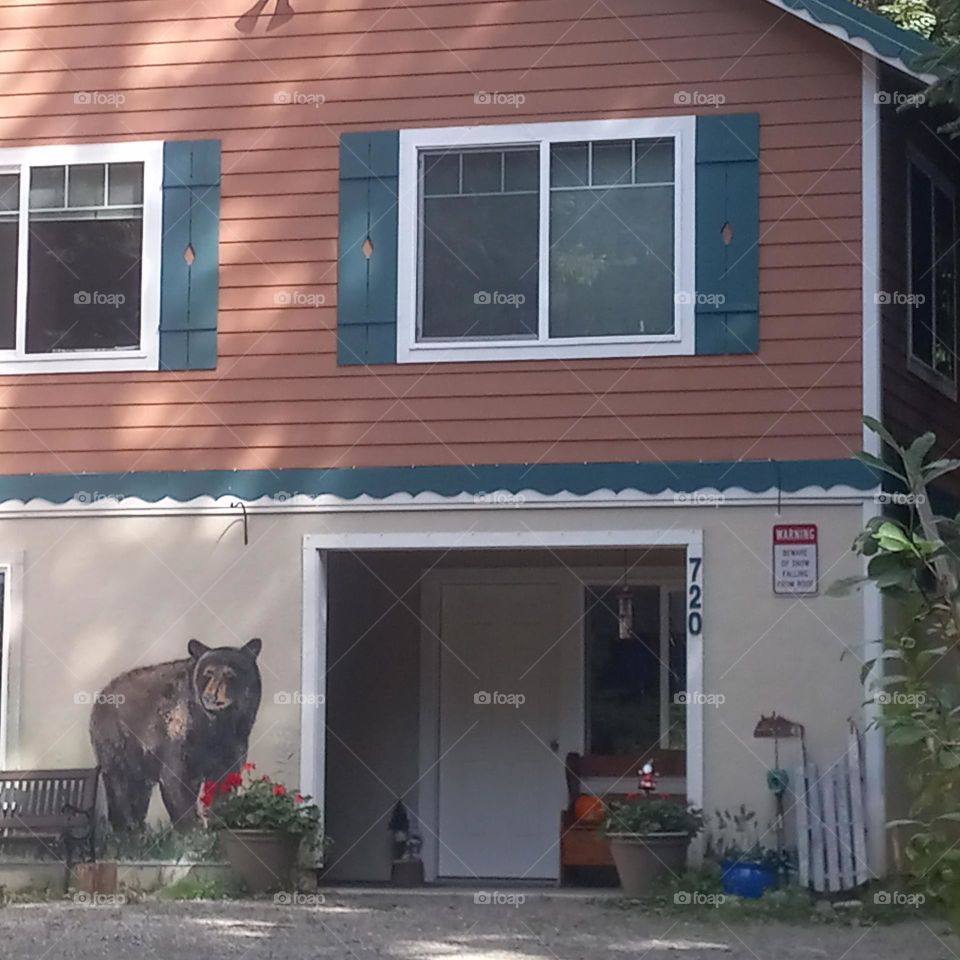 A garage entrance in a rural area with a hand painted scene including planters with flowers and greenery, fishing poles and focusing on a bear with quaint decorations of green shutters, benches and a white picket fence.