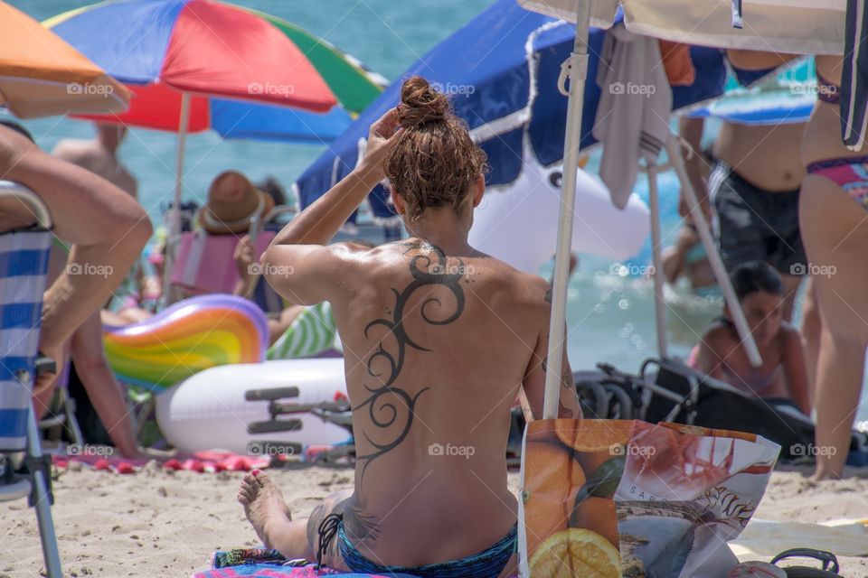 A woman sitting on the beach redoing her hair, on the back a beautiful tattoo