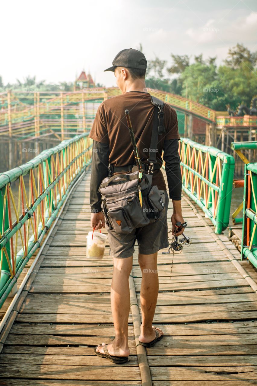 An angler is walking on a wooden bridge carrying fishing equipment