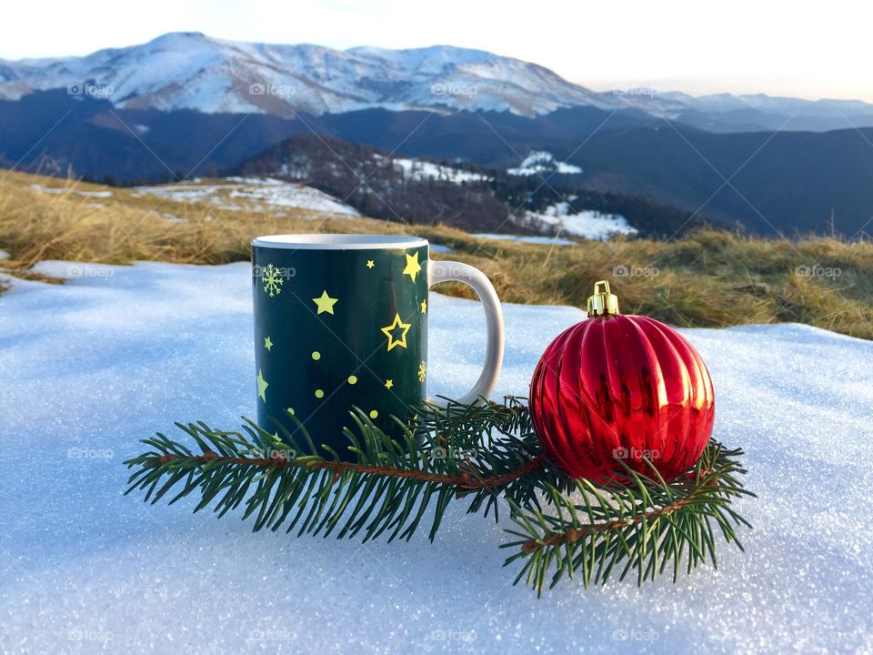 Snowflakes Christmas mug placed on snow with red globes and evegreen branches beside and snowy mountains in the background