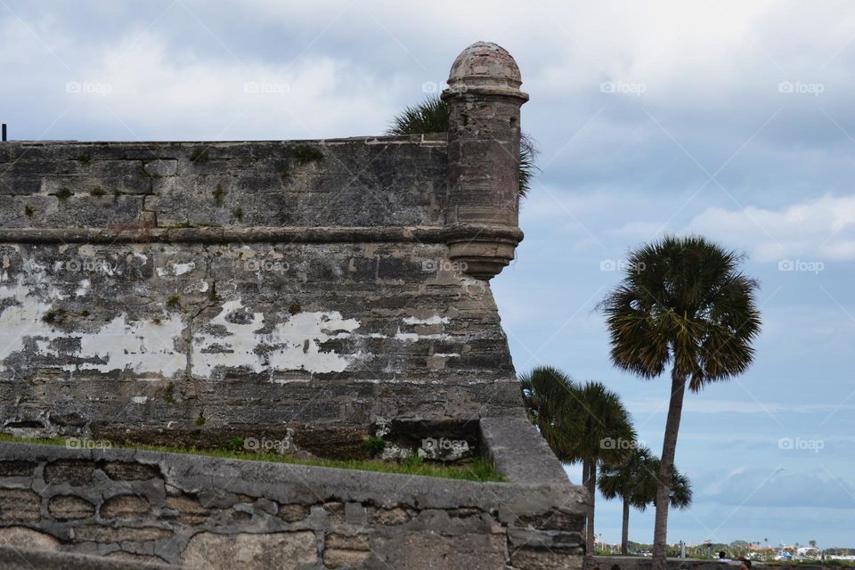 The corner of an old rock fort with a turret on top and palm trees next to it against a blue cloudy sky