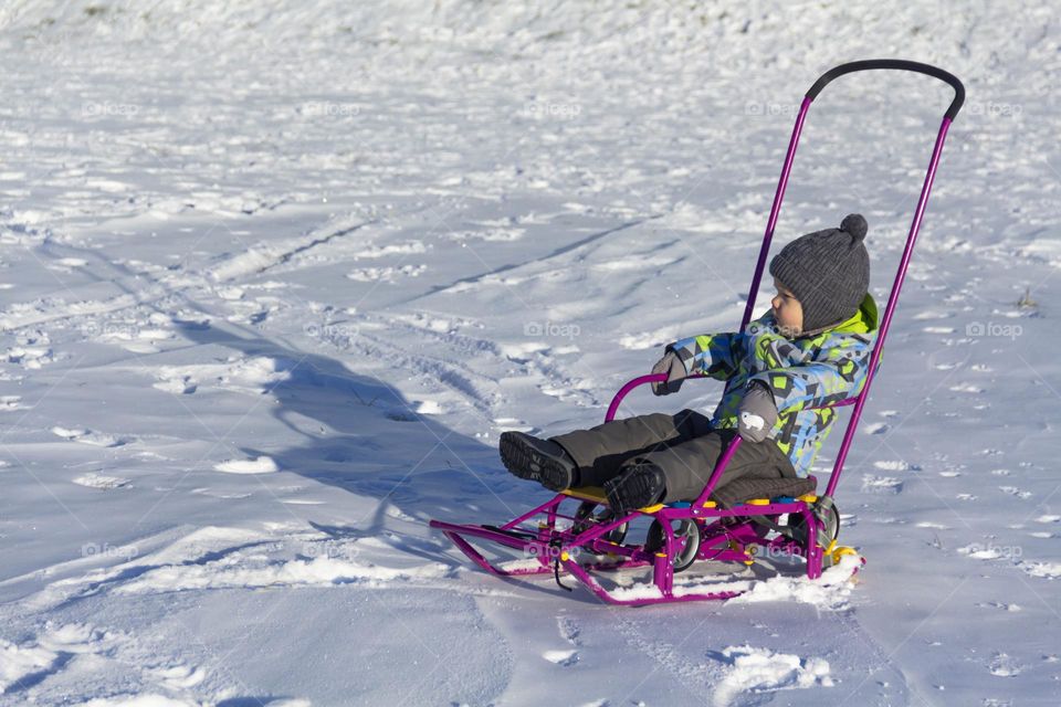 A child with a serious expression on his face in winter clothes jackets, pants, hat and boots in winter on white snow on the street and in the park in nature sledding and playing winter fun.