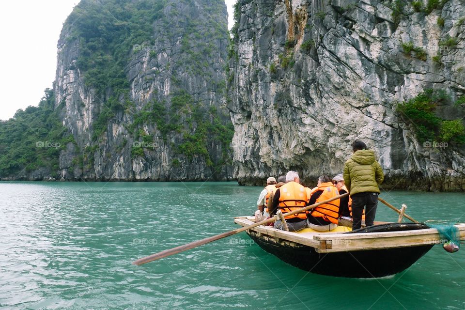 A boat is carrying tourists wearing orange life jackets, talking while watching the scenery.Shaggy dark gray rocky mountains, lots of trees.