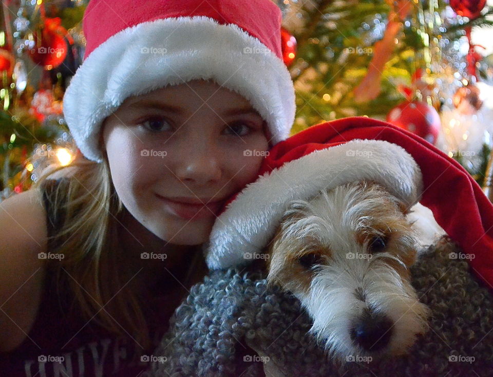 Girl and a dog with santa hats in front of a christmas tree