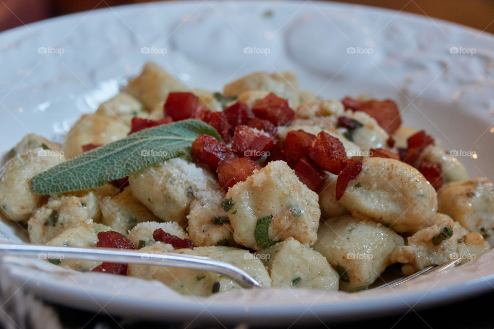 A close-up of a fresh plate of herbed gnocchi with chopped bacon and fresh sage in a butter sauce, plated on a white country plate.