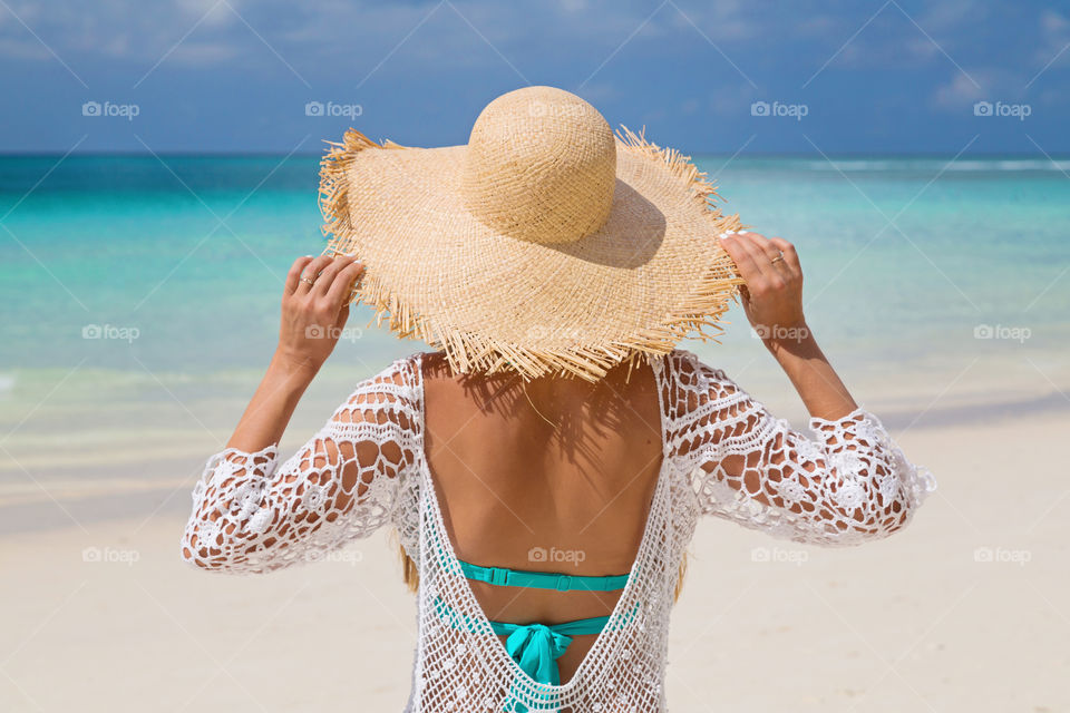 Slim young woman in bikini, beach tunica and big straw hat from behind standing near sea