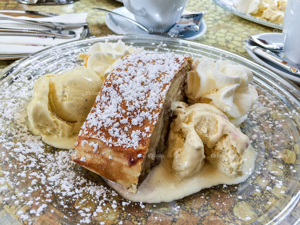 Café table with coffee cups and a glass plate with delicious Apfelstrudel with icecream and whipped cream outdoors in Germany in summer